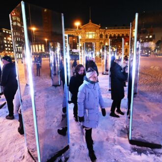 On a snowy, dark square, people walk around an art installation made of pillars that include mirrors and lights.