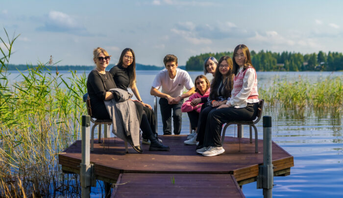 Half a dozen people of various nationalities are sitting on a dock in front of a scenic lake.