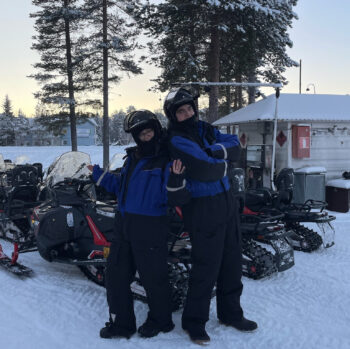 Two people wearing winter overalls and helmets pose in a snowy landscape in front of a row of snowmobiles.