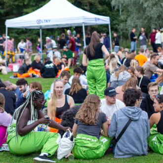 Groups of young people are sitting on a lawn in a park eating, drinking and talking.