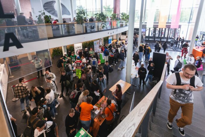 In the airy foyer of a campus building, students are conversing beside various tables stacked with brochures and merchandise.