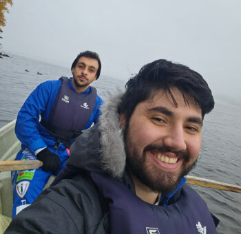 In a smiling selfie, two men are sitting in a rowboat on a lake.