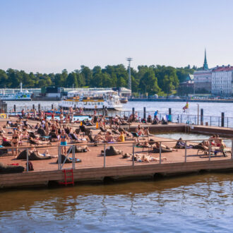 Ein Holzdeck, einschließlich eines Poolbeckens, schwimmt in einem Hafen mit Stadtgebäuden im Hintergrund.