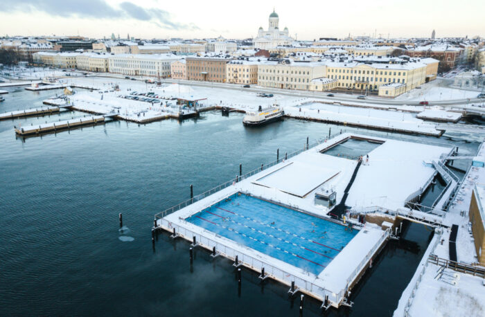 Um deck coberto de neve, incluindo várias bacias de piscina, flutua em um porto com prédios da cidade ao fundo.