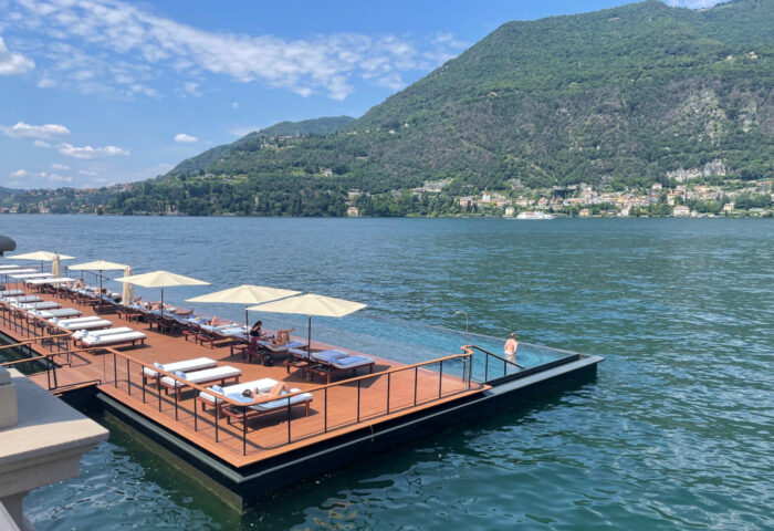 Um deck de madeira e uma bacia de piscina anexa flutuam em um lago com uma montanha ao fundo.