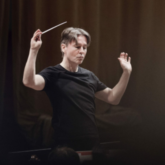 A man in a black T-shirt in front of a black background has his hands raised, with an orchestra conductor's baton in one hand.