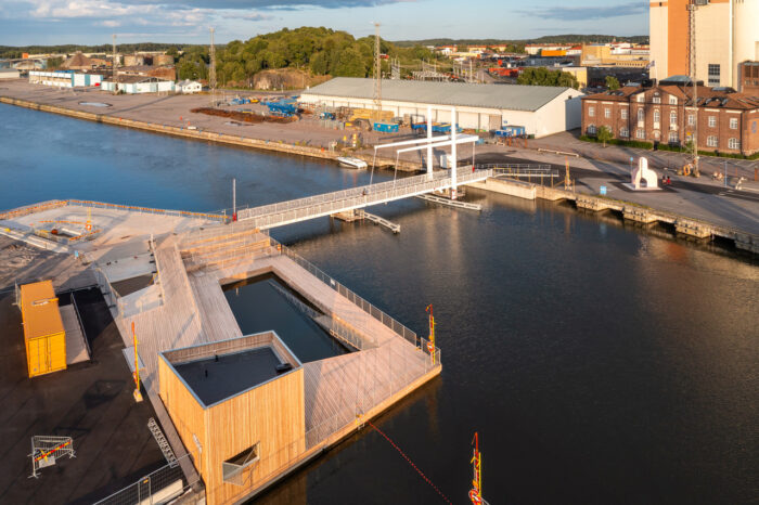 A partially constructed wooden deck, including a pool basin, floats on a harbour with city buildings in the background.