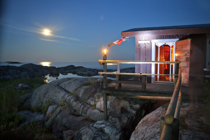 The door of a cottage, which is perched on beach rocks by the sea, is open as the moon shines through the clouds.