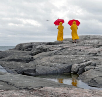 Duas mulheres com casacos amarelos longos seguram guarda-chuvas vermelhos, de pé sobre um rochedo, olhando para o mar.