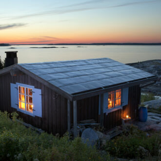 Light shines through the windows of a small cottage, situated by the sea, in evening dusk.