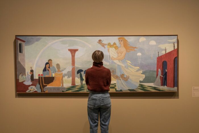 A woman is looking at a large painting that shows several people sitting at a table on a seaside terrace, with a rainbow, crashing waves and three parachutes visible in the background.