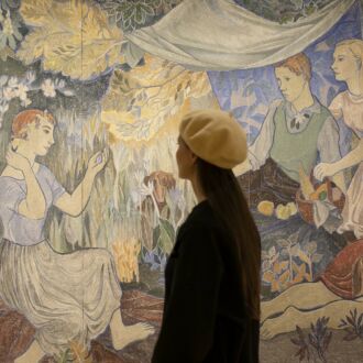 A woman in a beret is looking at a painting where people are picnicking in a forest and one person is playing a violin.