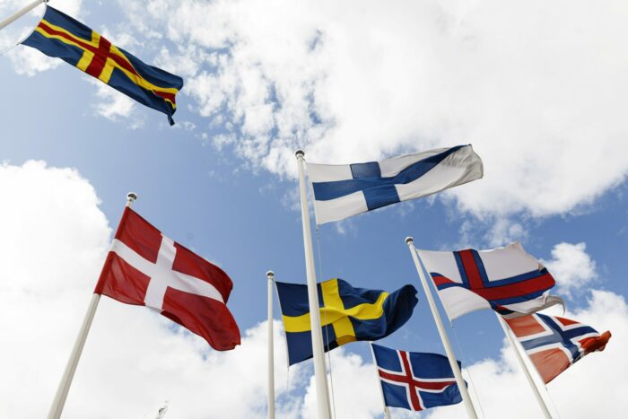 Seven national flags, each with a cross-shaped design, wave in the wind against in front of the sky.