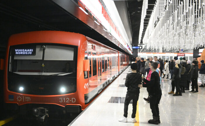 Um trem laranja de metrô está parado em uma estação iluminada decorativamente, com passageiros esperando na plataforma.