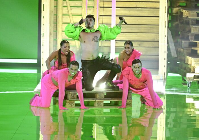 Un chanteur en blouson vert clair est à genoux sur une scène au milieu de danseurs et danseuses en tenue rose.