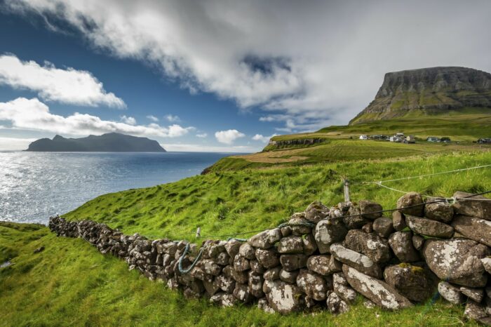 Under a blue sky with scattered clouds, a stone wall divides a grassy hillside overlooking a coastal village and distant island.