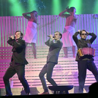 Cantores masculinos em um palco estão se apresentando com homens vestindo toalhas em frente a um cenário de madeira sob luzes verdes e roxas.