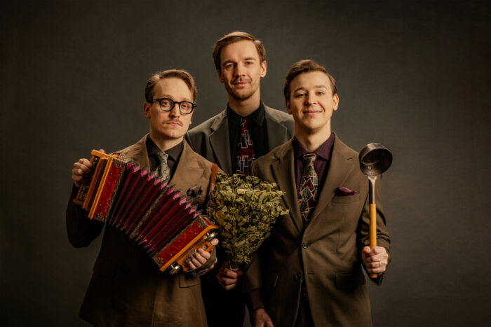 Trois hommes en costume prennent la pose devant l’objectif du photographe, l’un portant un accordéon en bandoulière, l’autre tenant un bouquet composé de branchettes feuillues, tandis que le troisième brandit une louche à long manche en bois. 