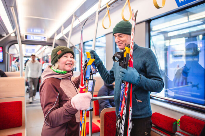 A man and a woman wearing winter hats and sporty clothing and holding skis ride a tram.