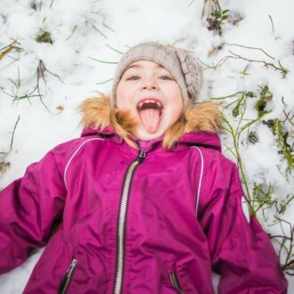 A young child in a winter coat lies in the snow with their mouth open, smiling and sticking their tongue out.