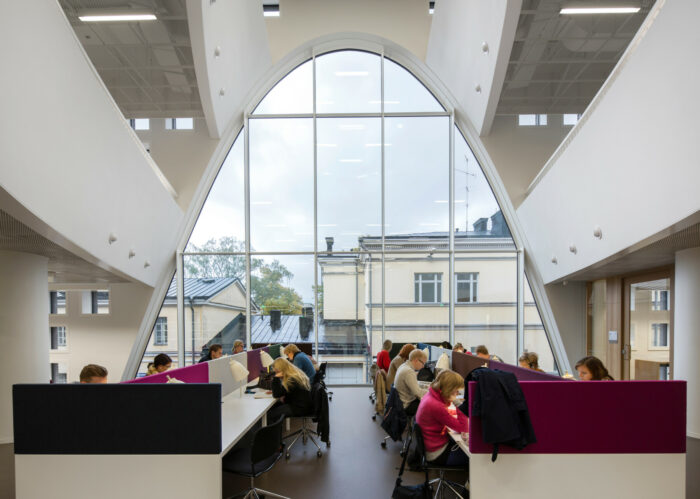 Des gens assis à des tables travaillent dans des espaces compartimentés au milieu d’un vaste open space lumineux et moderne surmonté d’une haute baie vitrée en ogive. 