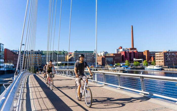 Several cyclists rides across a bridge on a sunny day, with buildings visible in the background.