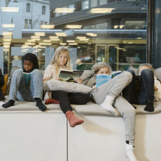 On a sofa in front of a large glass window looking out to a city street, eight children sit and read.