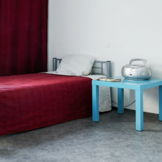Un dormitorio pulcramente ordenado, con una cama cubierta por una colcha de color rojo oscuro y una mesilla de noche azul claro sobre la que hay radio.