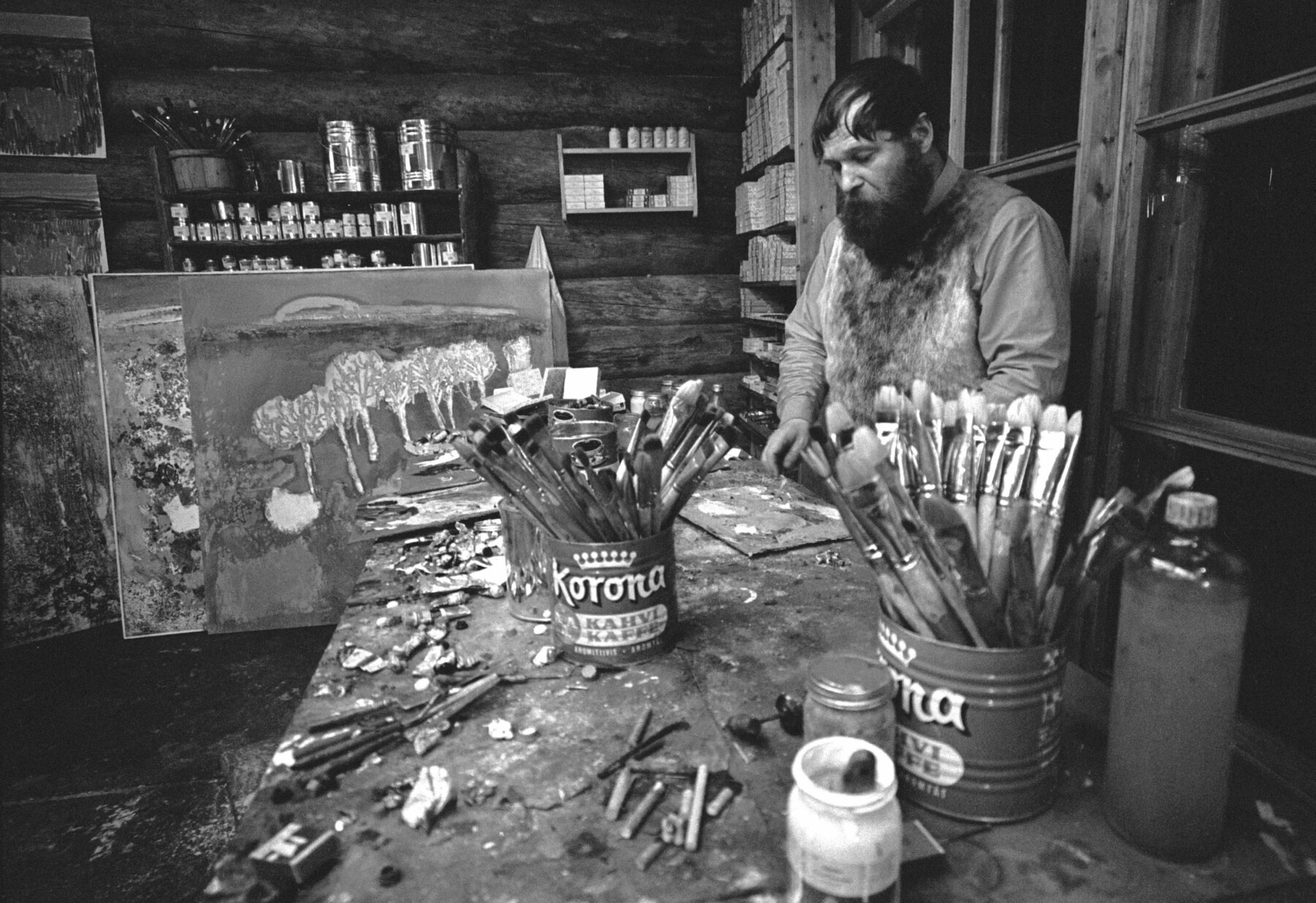 A man with a beard is working in an art studio, with brushes and other supplies on a table.