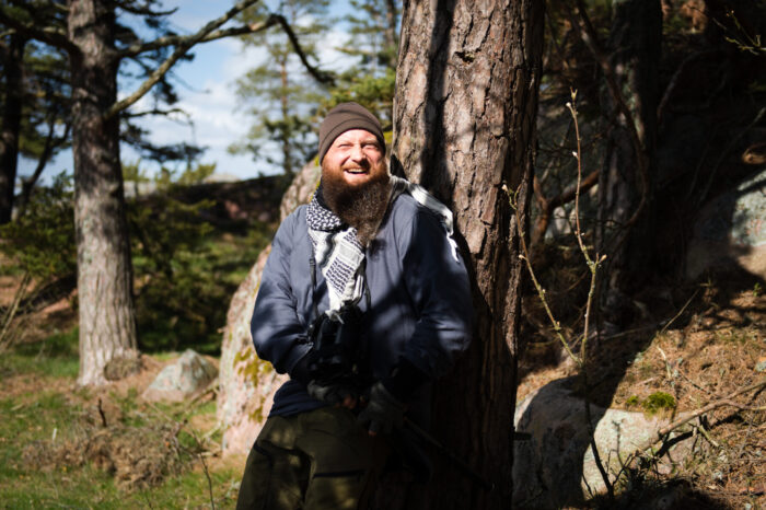 A man dressed for cold weather leans against a pine tree, laughing with sunlight warming his face.