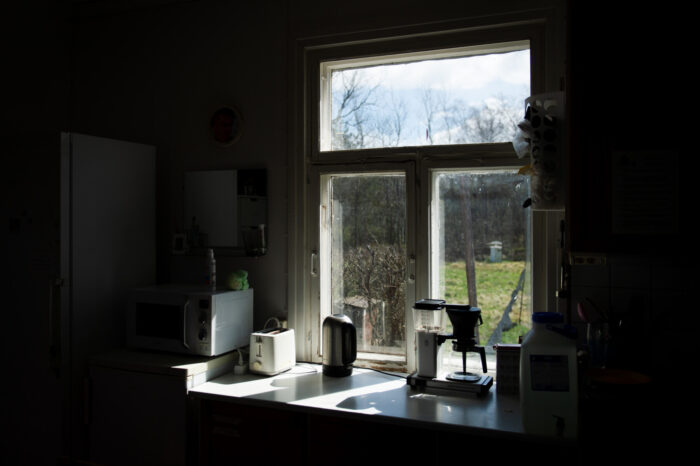 Inside the dark interior of a cottage, a bright kitchen window frames the sunlit grass and sky outside.