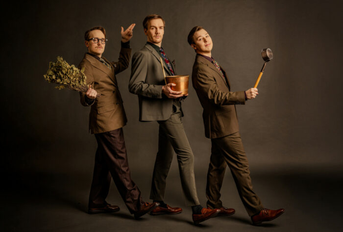 Three men in suits stand in a row, each with a piece of sauna equipment: a leafy bouquet of branches, a metal bucket, and a ladle.