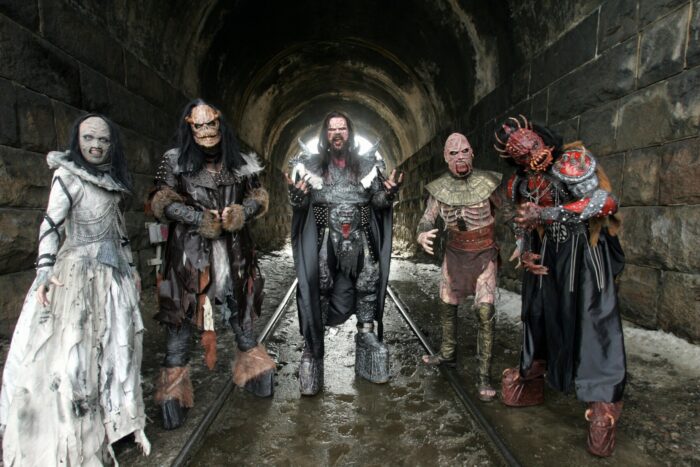 Five people in fearsome monster costumes are standing in a row in a stone underpass.