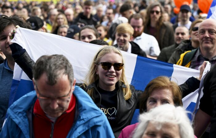 In a crowd of people, one woman is holding up a large Finnish flag.