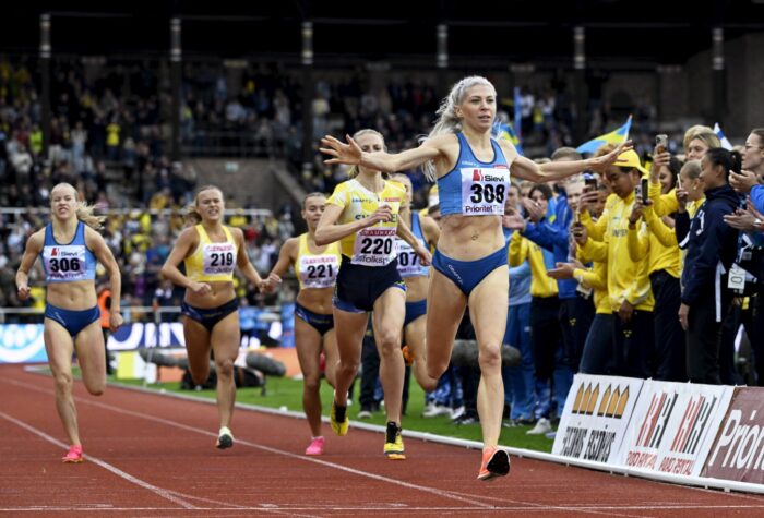 A Finnish runner crosses the finish line in front of four other competitors. Photographers line the track and spectators are in the background.