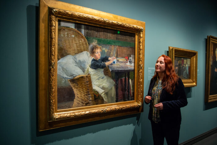 A woman looks at a large painting on a gallery wall, showing a child sitting in a wicker chair.