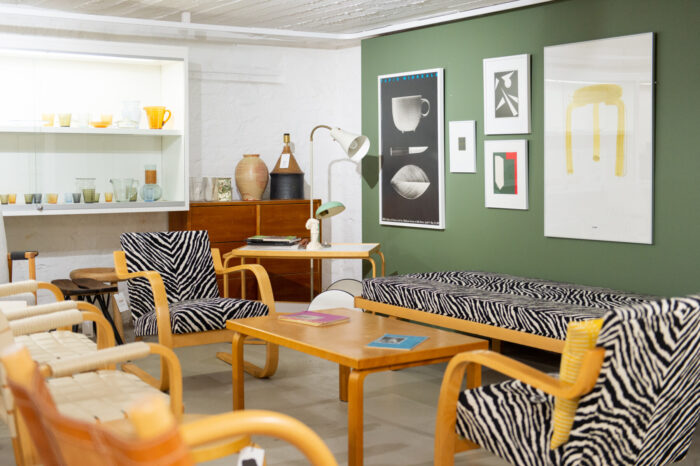 Différents articles sont présentés dans un espace aménagé au sein d’un magasin de mobilier, dont des chaises, de la verrerie et des tableaux accrochés à un mur vert.
