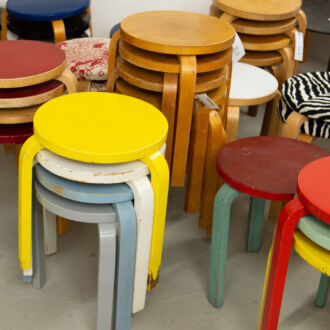 Banquetas redondas de madeira, com três pernas e cores variadas, dispostas em pilhas de quatro ou cinco em uma loja de móveis.
