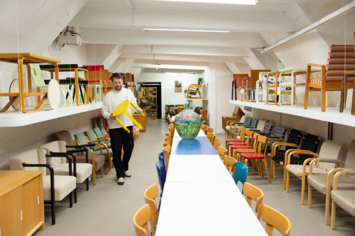 Dans un magasin de meubles, un homme portant un tabouret jaune passe devant une longue table flanquée de chaises et d’autres pièces de mobilier.