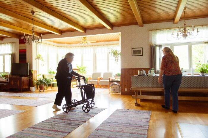 An elderly woman walks through a living room using a walker.