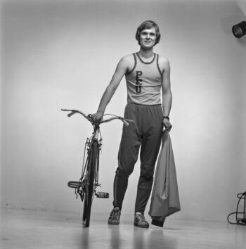 A man in a tracksuit poses with one hand resting on a bicycle and the other hand holding a jacket.
