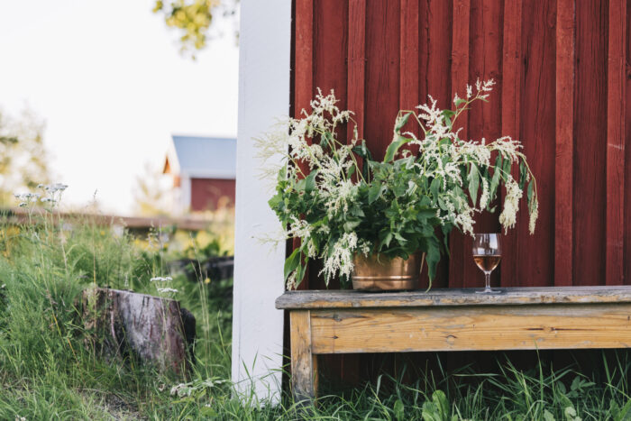 Em um banco ao ar livre, ao lado de um muro vermelho, há um buquê de flores silvestres e uma taça de vinho. Ao fundo, um celeiro vermelho.