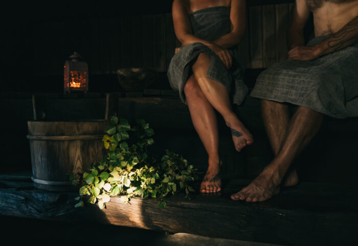 Um homem e uma mulher sentam-se em uma sauna mal iluminada com uma lanterna, um balde de madeira e um feixe de galhos de bétula.