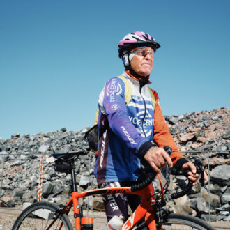 An older man in athletic cycling gear walks his bicycle along a stone embankment.