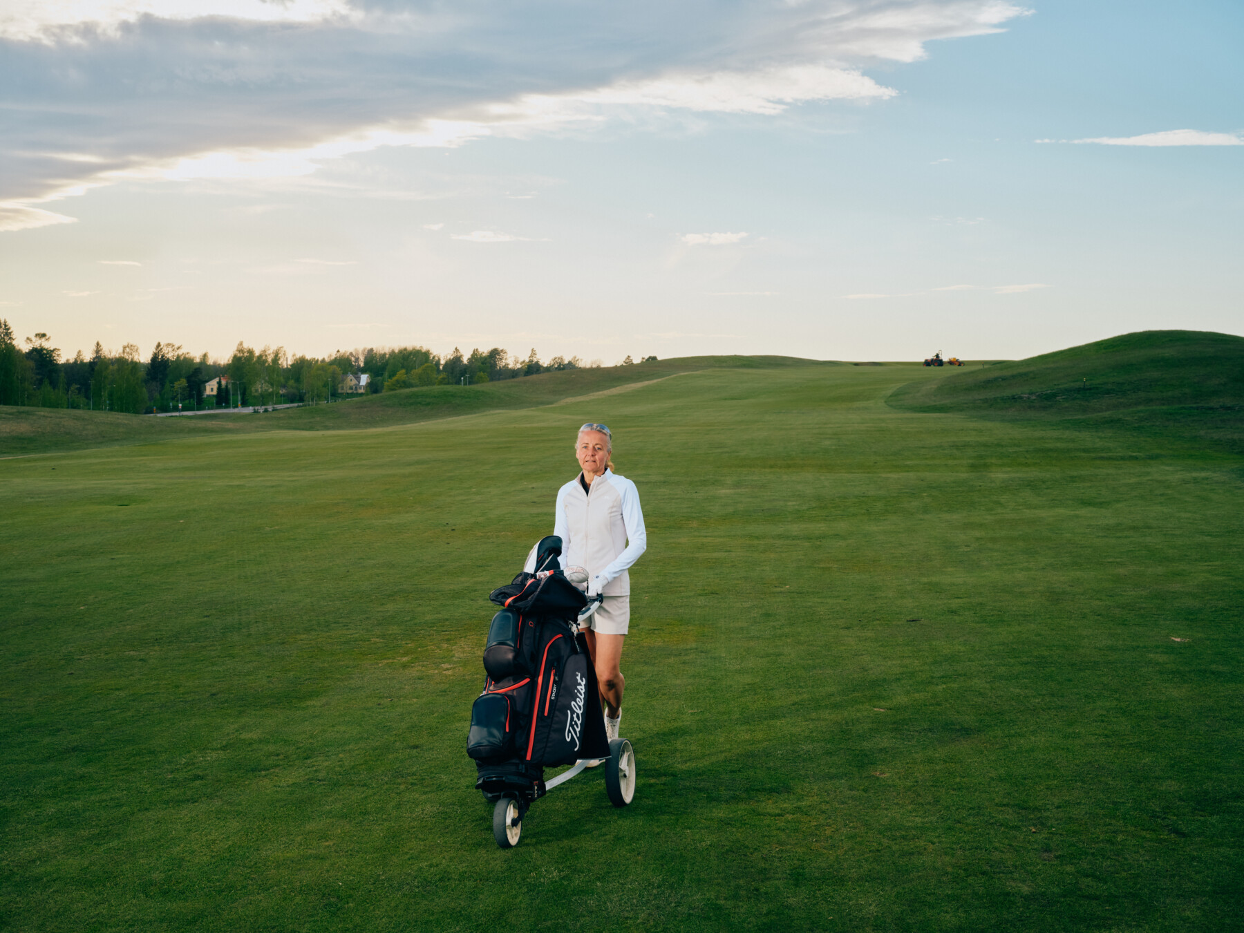 A woman stands on a golf course with a golf bag.
