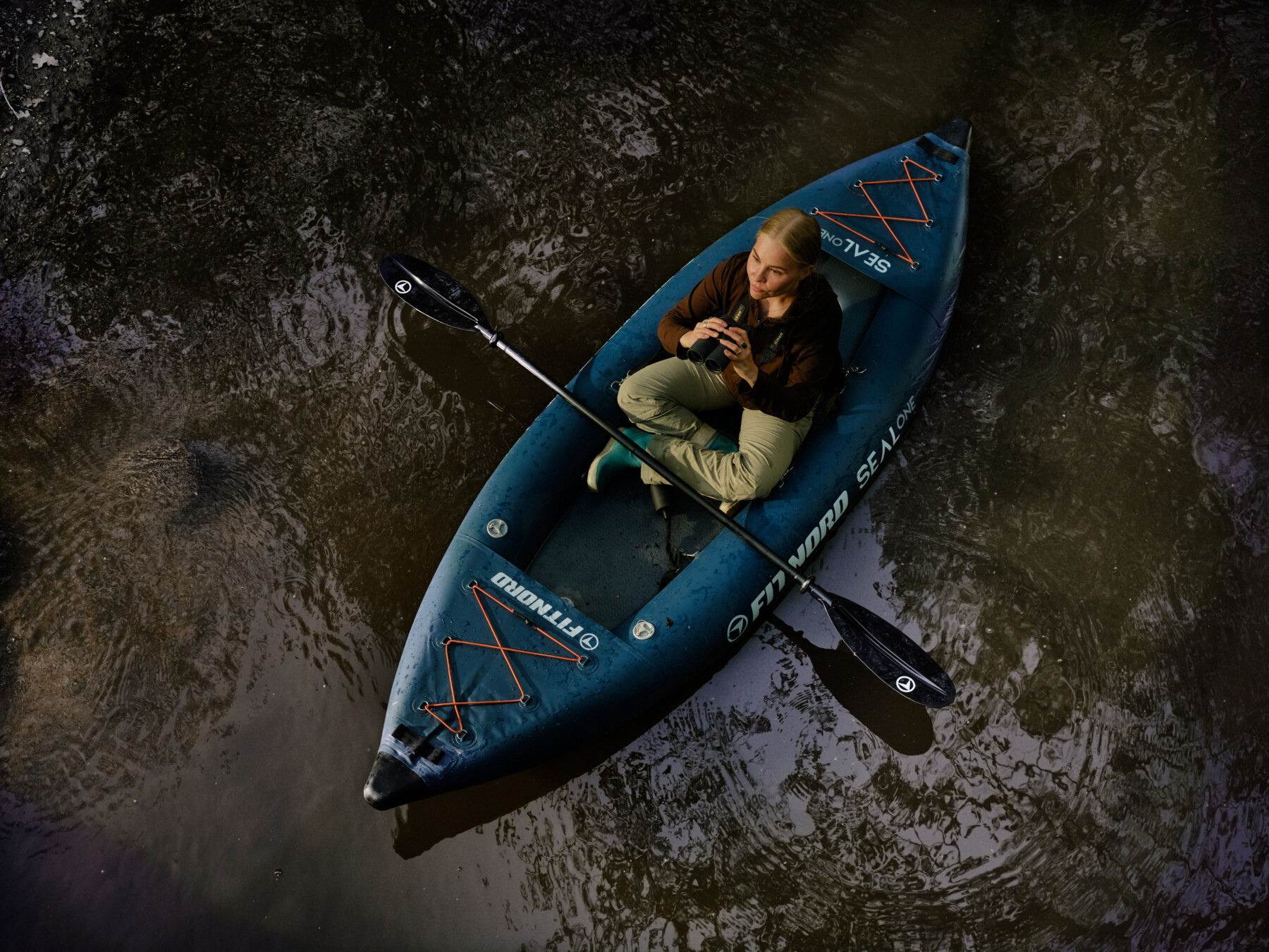 A woman sits on a kayak holding binoculars.