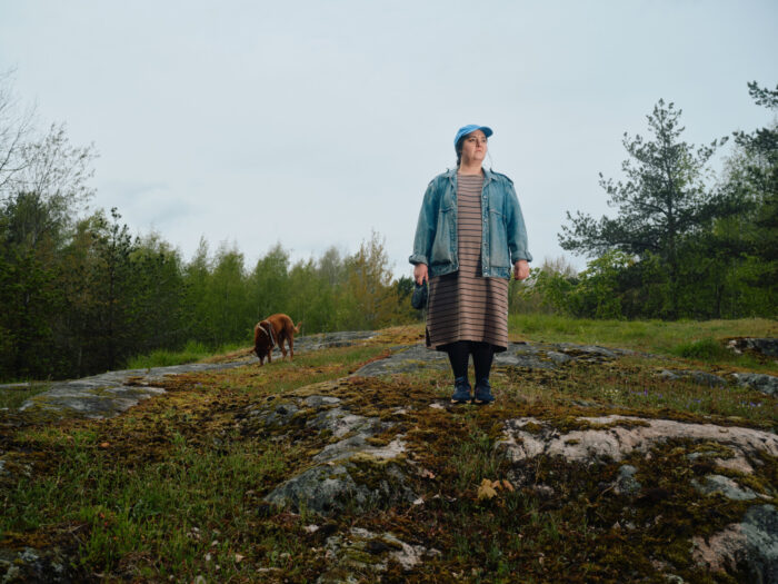 Une femme accompagnée d’un chien se tient debout sur une colline rocailleuse entourée d’espaces boisés. 