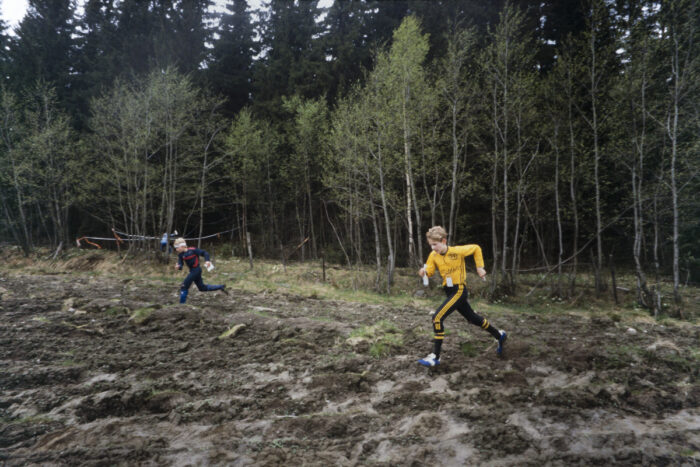 Two people run across a muddy field during an orienteering event, with a forest in the background.