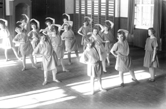 A women’s gymnastics team practices indoors in an old black-and-white photo, wearing period-appropriate, loose-fitting exercise outfits.