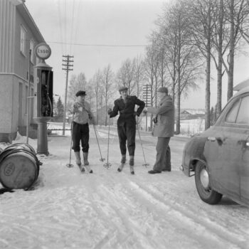 Two men with skis stand in front of a gas station during winter while another man without skis stands nearby.